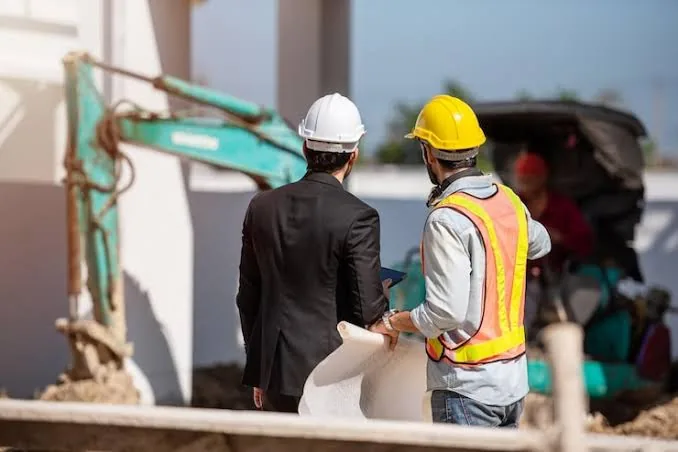 Tecnico con casco e gilet ad alta visibilità e responsabile di cantiere osservano l’area durante i lavori, con escavatore sullo sfondo
