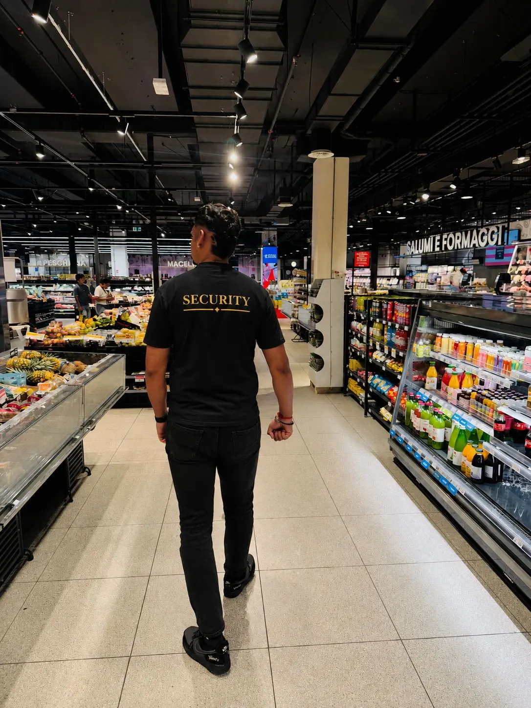 Operatore OnTopService di spalle, con maglia SECURITY, mentre percorre la corsia di un supermercato accanto ai banchi freschi